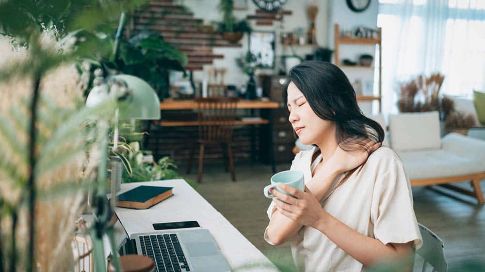 Lady sitting at desk at home, clutching her neck with her hand suggesting discomfort, whilst holding a mug in the other hand.